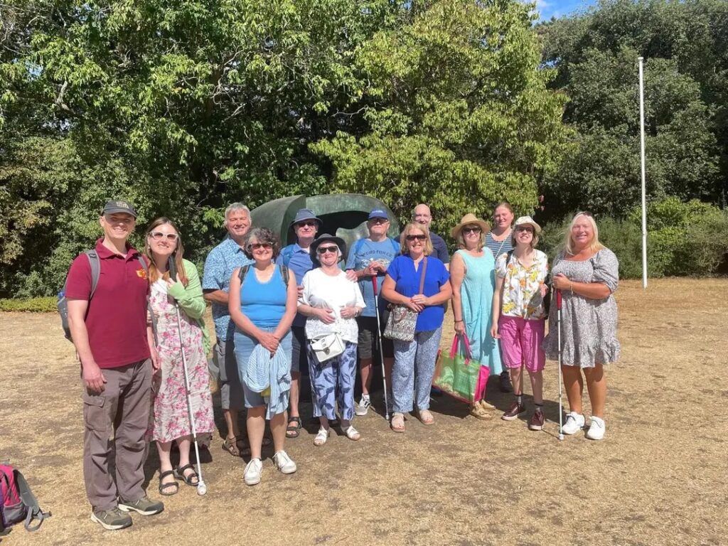 Photograph of Bats members Usb, Becky, Mike, Cheryl, Pete M, Sue, Bill, Berys, Shaun, Rose, Linda, Kayleigh and Ali all about to go into Kew Gardens, in the August Sunshine with a lovely green background of plants behind them all.